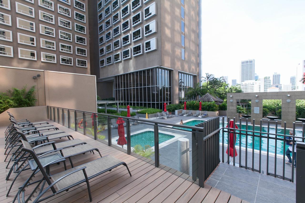 新加坡卡尔登酒店【Sg清洁认证】 外观 照片 The photo shows a modern outdoor area of a building featuring a swimming pool. In the foreground, there are several lounge chairs arranged on a wooden deck. Behind the chairs, there are two small swimming pools, which appear to be surrounded by green